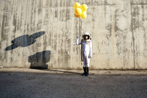 Girl wearing space suit holding yellow balloon standing against wall during sunny day - GGGF00010