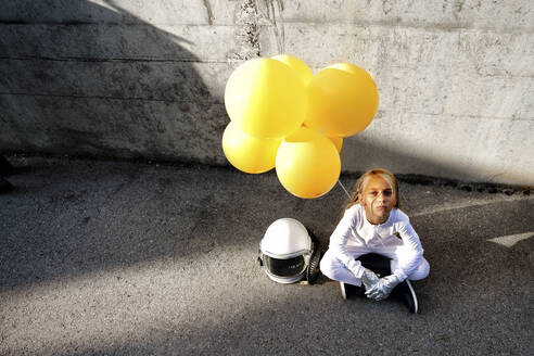 High angle view of girl holding yellow balloon sitting against wall during sunny day - GGGF00011
