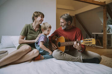 Vater spielt Gitarre, während Mutter und Sohn auf dem Bett im Schlafzimmer sitzen - MFF06622