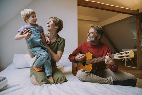 Father playing guitar while mother and son enjoying music while sitting on bed in bedroom at home stock photo