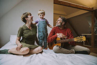 Sohn steht zwischen Mutter und Vater und spielt Gitarre, die zu Hause im Schlafzimmer sitzen - MFF06624