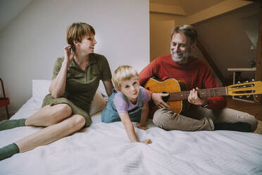 Happy father playing guitar while mother and son sitting on bed at home - MFF06625