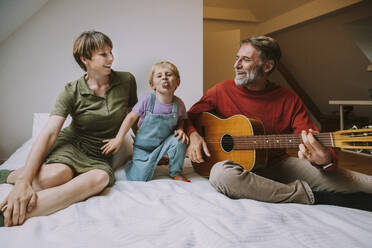 Sohn streckt die Zunge heraus, während der Vater Gitarre spielt und die Mutter neben ihm auf dem Bett sitzt - MFF06626