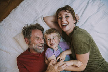 Smiling mother, father and son embracing each other while lying on bed at home - MFF06632