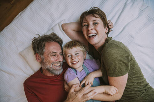 Lächelnde Mutter, Vater und Sohn, die sich gegenseitig umarmen, während sie zu Hause auf dem Bett liegen - MFF06632