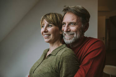 Smiling couple looking away embracing while standing at home - MFF06636