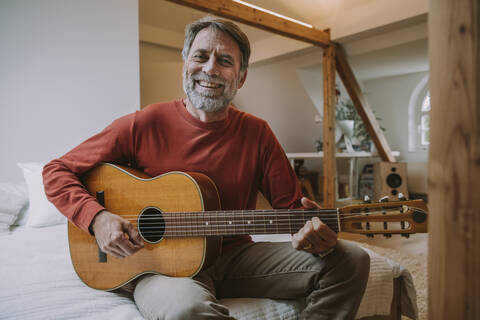 Mature man playing guitar while sitting on bed in bedroom at home stock photo