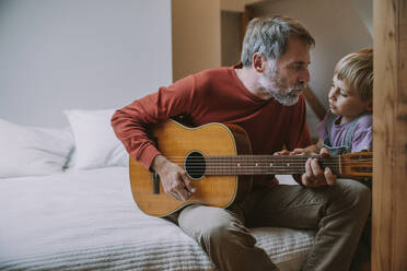 Vater spielt Gitarre vor seinem Sohn, während er zu Hause auf dem Bett sitzt - MFF06642