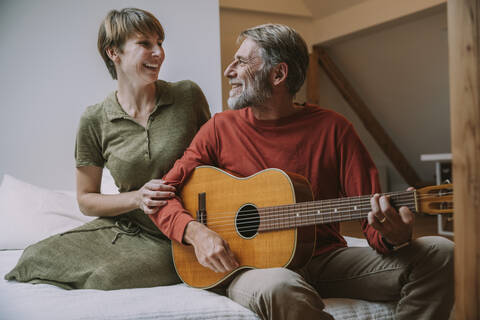 Mature man playing guitar while woman looking at him sitting on bed in bedroom stock photo