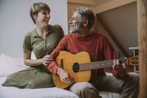 Reifer Mann spielt Gitarre, während die Frau ihn im Schlafzimmer auf dem Bett sitzend anschaut - MFF06643