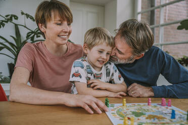 Vater kuschelt mit seinem Sohn, während die Mutter daneben sitzt und zu Hause ein Brettspiel spielt - MFF06658