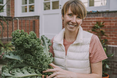 Lächelnde erwachsene Frau, die ein Grünkohlblatt hält, während sie im Hinterhof steht - MFF06669