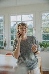 Smiling woman talking on smart phone looking away while standing at home - MFF06691