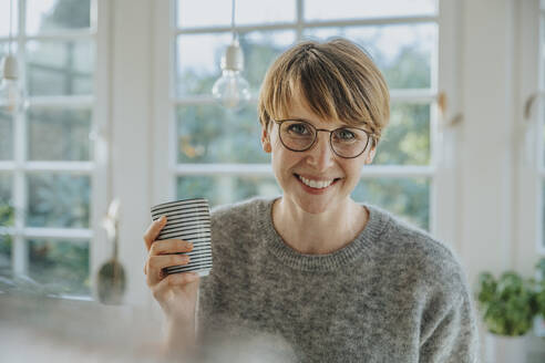 Smiling mid adult woman holding tea cup while standing at home - MFF06694