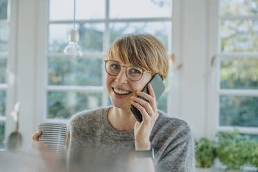 Lächelnde Frau, die mit ihrem Smartphone telefoniert und eine Teetasse hält, während sie zu Hause steht - MFF06695