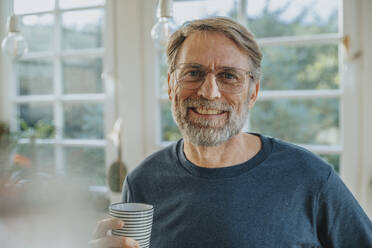 Smiling mature man with tea cup standing at home - MFF06697