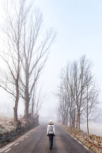 Rückenansicht einer anonymen Wanderin in warmer Kleidung, die auf einer leeren Asphaltstraße zwischen blattlosen Bäumen an einem kalten, nebligen Tag auf dem Land in Bellver de Cerdaña, Katalonien, wandert - ADSF17019