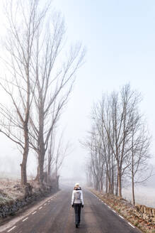 Back view of anonymous female hiker in warm clothes walking on empty asphalt road among leafless trees in cold misty day in countryside in Bellver de Cerdaña , Catalunya - ADSF17019