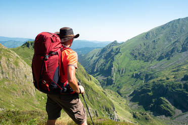 Rückenansicht eines nicht erkennbaren männlichen Wanderers mit Rucksack und Trekkingstock, der auf einer grasbewachsenen Bergkuppe steht und die spektakuläre Kulisse eines Bergkamms an einem Sommertag bewundert - ADSF17024