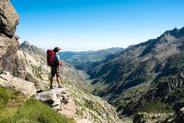 Back view of unrecognizable male hiker with backpack and trekking stick standing on rocky hilltop and admiring spectacular scenery of mountain ridge in summer day - ADSF17025