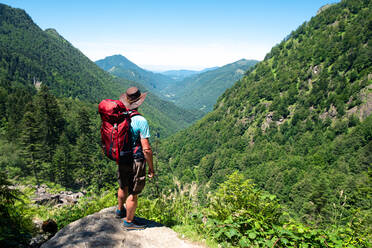 Back view of unrecognizable male hiker with backpack and trekking stick standing on grassy hilltop and admiring spectacular scenery of mountain ridge in summer day - ADSF17026