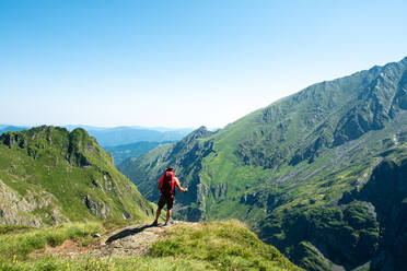 Back view of distant unrecognizable male hiker with backpack and trekking stick standing on grassy hilltop and admiring spectacular scenery of mountain ridge in summer day - ADSF17027