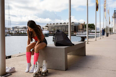 Slim woman in summer wear sitting on bench in city and preparing for roller skating - ADSF17065