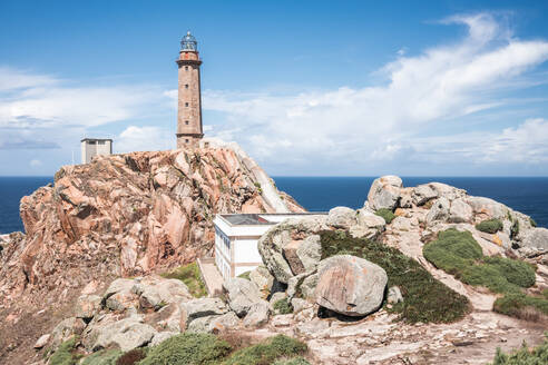 Malerische Landschaft mit altem Leuchtturm und Häusern auf einer rauen Felsklippe in der Nähe des Meeres auf der Halbinsel Kap Vilan an der spanischen Küste gegen einen bewölkten blauen Himmel an einem Sommertag - ADSF17071