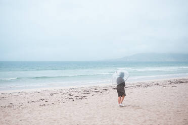 Seitenansicht eines nicht erkennbaren Reisenden in einem Regenmantel mit einem durchsichtigen Regenschirm, der auf einem leeren Sandstrand in Richtung des wogenden Meeres an einem bewölkten Tag spazieren geht - ADSF17074