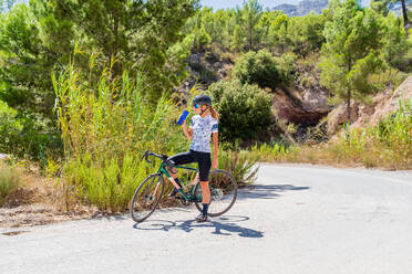 Full body of fit female bicyclist in sportswear and helmet drinking fresh water from bottle while resting after riding bike up mountain road - ADSF17085