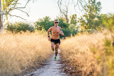 Hemdloser männlicher Sportler mit Sonnenbrille, der auf einem schmalen Fußweg inmitten von hohem Gras während eines Trainings in der Natur läuft - ADSF17090