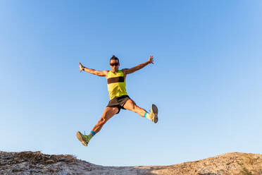 Von unten Ganzkörperaufnahme eines athletischen Mannes in Sportkleidung und Sonnenbrille, der mit ausgestreckten Armen gegen den wolkenlosen blauen Himmel hochspringt - ADSF17092