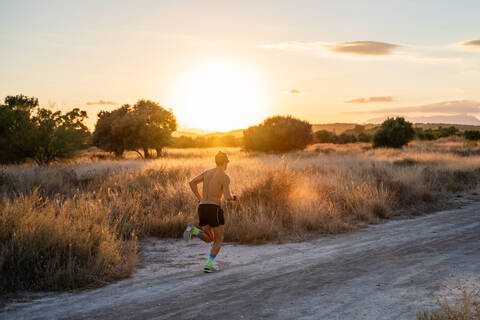 Rückenansicht eines nicht erkennbaren aktiven männlichen Sportlers mit kräftigem Körper, der eine Landstraße entlangläuft und während eines intensiven Trainings im sommerlichen Sonnenuntergang die Zeit auf seiner Armbanduhr überprüft, lizenzfreies Stockfoto