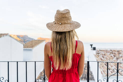 Rückenansicht einer nicht wiedererkennbaren Touristin, die mit Strohhut in erhobener Hand auf der Terrasse steht und den Sommerurlaub genießt, während sie das erstaunliche Stadtbild bewundert - ADSF17104