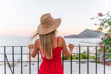 Back view of unrecognizable female tourist standing with straw hat in raised hand on terrace and enjoying summer vacation while admiring amazing cityscape - ADSF17106