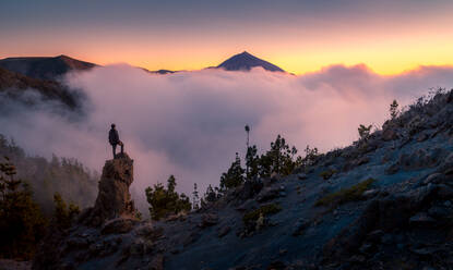 Back view of anonymous explorer standing on rock and observing majestic scenery of mountains under thick fog in morning during vacation in Tenerife - ADSF17122