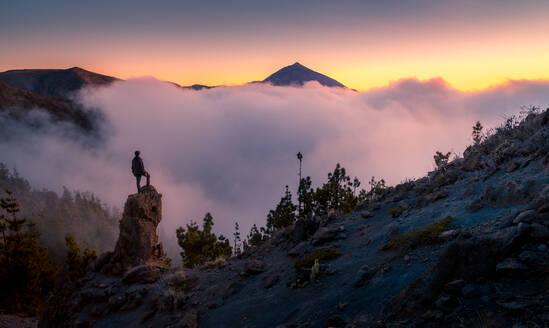 Rückansicht eines anonymen Forschers, der auf einem Felsen steht und die majestätische Berglandschaft unter dichtem Nebel am Morgen während des Urlaubs auf Teneriffa beobachtet - ADSF17122