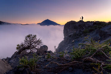 Seitenansicht eines anonymen Fotografen, der auf einem Felsen steht und die majestätische Berglandschaft unter dichtem Nebel am Morgen während seines Urlaubs auf Teneriffa beobachtet - ADSF17123