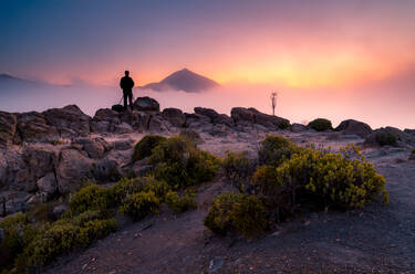 Rückansicht eines anonymen Fotografen, der auf einem Felsen steht und die majestätische Berglandschaft unter dichtem Nebel am Morgen während seines Urlaubs auf Teneriffa beobachtet - ADSF17124