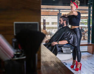 Seitenansicht einer stilvollen Meisterin in schwarzer Lederhose und roten Stöckelschuhen, die die Haare eines bärtigen männlichen Kunden bei der Arbeit im Friseurladen trocknet - ADSF17143