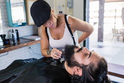 Seitenansicht einer stilvoll tätowierten Kundin, die sich mit einer elektrischen Maschine einen Bart rasiert, während sie in einem professionellen Friseurladen arbeitet - ADSF17145