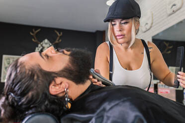 Seitenansicht einer stilvoll tätowierten Kundin, die sich mit einer elektrischen Maschine einen Bart rasiert, während sie in einem professionellen Friseurladen arbeitet - ADSF17147