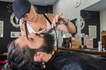 Seitenansicht einer stilvoll tätowierten Kundin, die sich mit einer elektrischen Maschine einen Bart rasiert, während sie in einem professionellen Friseurladen arbeitet - ADSF17148
