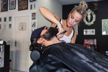 Seitenansicht einer stilvoll tätowierten, bärtigen Kundin, die sich mit einem Rasiermesser rasiert, während sie in einem professionellen Friseurladen arbeitet - ADSF17151