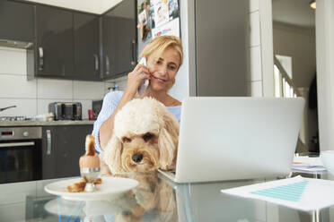 Reife Frau, die mit einem Smartphone spricht und mit einem Hund in der Küche zu Hause sitzt - PMF01426