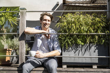 Lächelnder Mann mit Kaffeetasse auf dem Balkon an einem sonnigen Tag - UUF21852