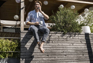 Mann schaut weg, während er auf dem Balkon sitzend mit seinem Smartphone spricht, während eines sonnigen Tages - UUF21855