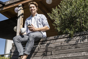 Mittlerer erwachsener Mann mit Smartphone und Kaffeetasse auf dem Balkon an einem sonnigen Tag - UUF21857