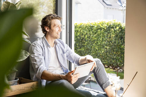 Lächelnder Mann, der ein digitales Tablet hält und wegschaut, während er zu Hause sitzt - UUF21858