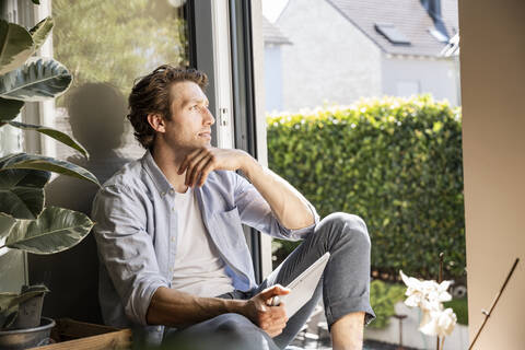 Man with hand on chin holding digital tablet looking away while sitting at home stock photo